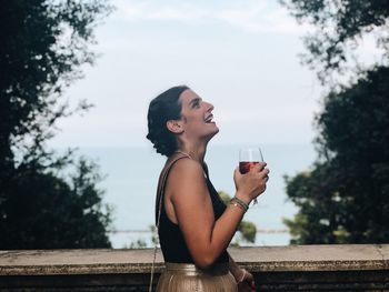 Side view of cheerful woman having wine while standing against sea