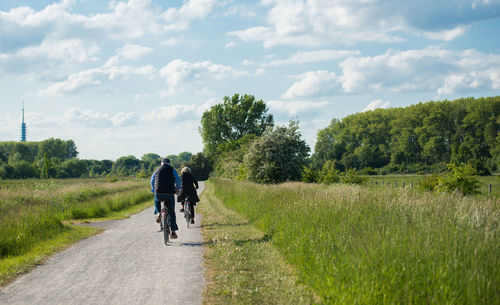 Rear view of man and woman riding bicycles on road amidst field against sky