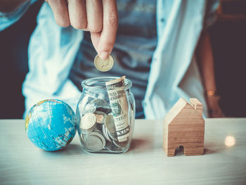 Close-up of hand holding coin over jar by globe and model home on table