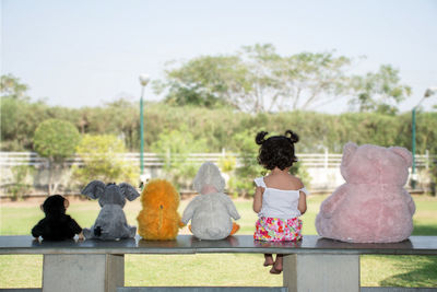 Rear view of girl sitting by stuffed toys on bench at park