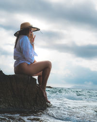 Full length of woman wearing hat sitting on rock against sea and sky