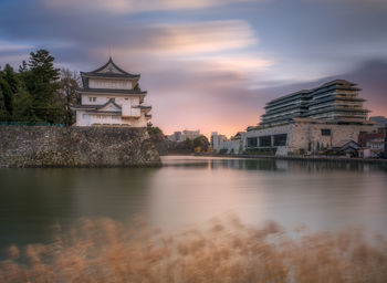 Buildings by river against sky during sunset