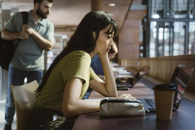 Focused young woman studying on laptop while leaning on elbow at desk in university library