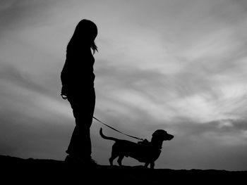 Side view of silhouette woman standing with dachshund on land against sky