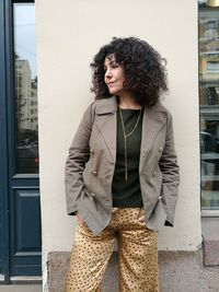 Mature woman with hands in pockets standing against wall