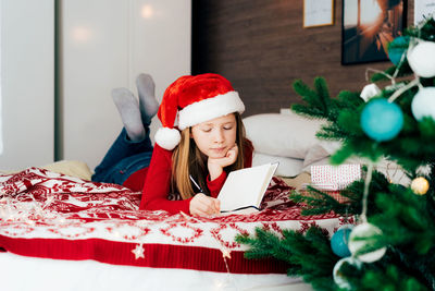A cute redhaired teen girl in a santa hat lying on the bed writes christmas wishes in a notebook.