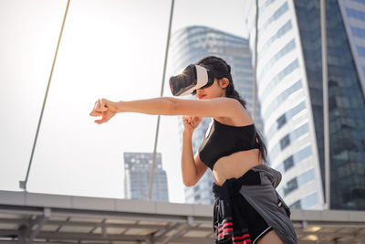 Young woman using virtual reality simulator punching against building in city