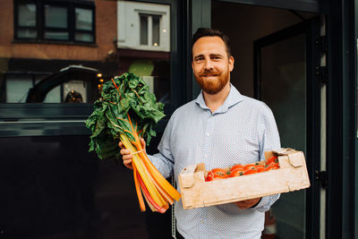 Delivery man holding box of tomatoes and greens at restaurant entrance