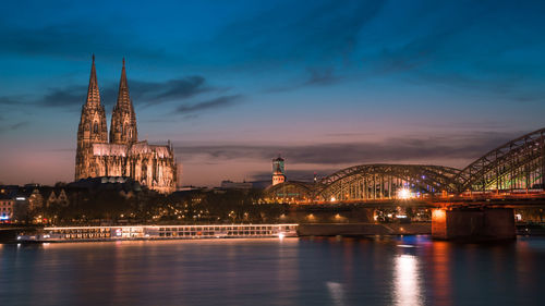 Illuminated bridge over river by buildings against sky at night