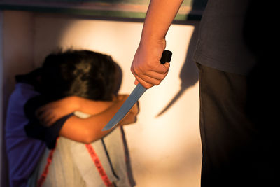 Midsection of criminal holding knife threatening scared woman sitting by wall