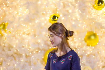 Young woman looking away while standing against illuminated christmas lights