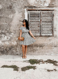 Front view of young woman in polka dot dress posing in front of weathered wall and old window.