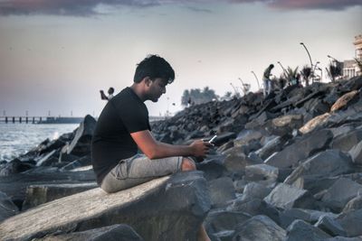 Side view of young man using smart phone on rock at beach against sky