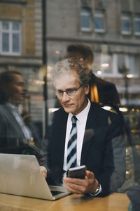 Smiling businessman with smart phone using laptop in cafe seen through glass window