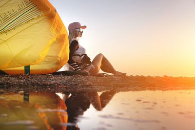 Attractive stylish young caucasian woman in cap sunglasses and kitesurfer outfit sits on the sandy