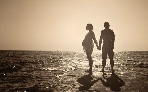 Silhouette couple holding hands while standing by sea against sky during sunset
