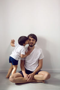 Father and son in white t-shirts and shorts sit at home against the wall