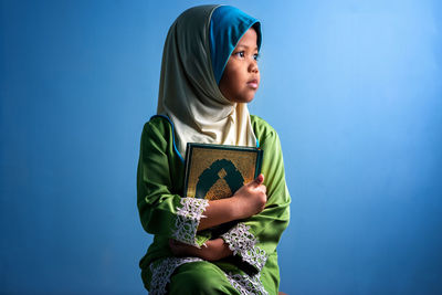 Girl wearing hijab holding koran while sitting against blue background