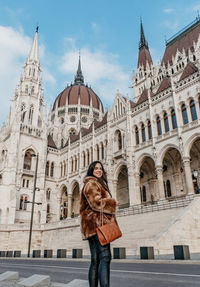 Young woman wearing stylish warm coat, standing in front of hungarian parliament in budapest hungary