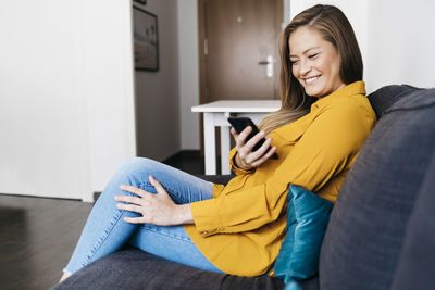 Smiling young woman using mobile phone while sitting on sofa at home