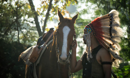 Side view of man wearing feathered headdress while holding horse in forest