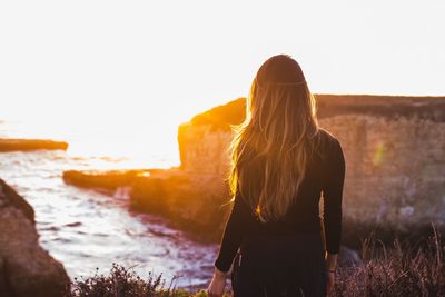 Rear view of standing by rock in sea against clear sky during sunset