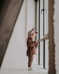 Toddler baby girl in funny hat with ears standing near the window