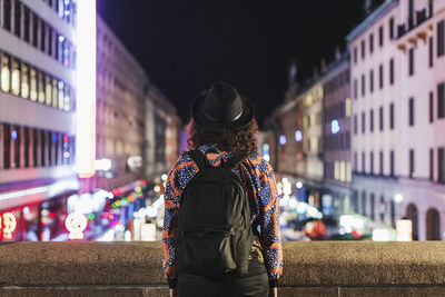 Rear view of woman carrying backpack standing on bridge in city at night