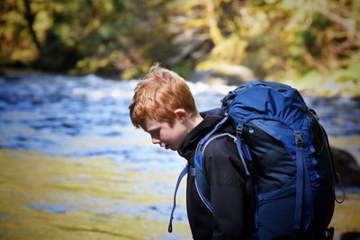 Side view of boy carrying backpack looking down while standing by river in forest