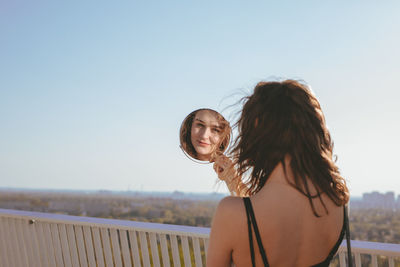 Rear view of young woman looking in mirror against clear sky