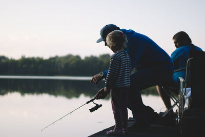 Rear view of daughter and father fishing in lake against sky