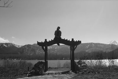 Man sitting on arbor at lakeshore against sky