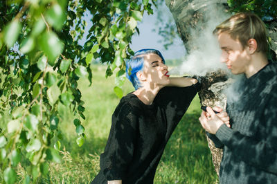 Young woman exhaling smoke on friend by tree