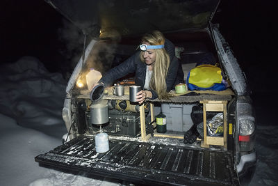 Smiling young woman preparing food in off-road car at night during winter