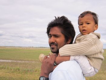 Portrait of father carrying son on shoulder while standing at field against sky