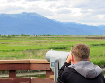 Rear view of boy looking through coin-operated binoculars on grassy field