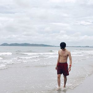 Shirtless young man walking on shore at beach against cloudy sky