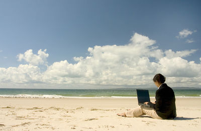 Rear view of mid adult woman using laptop while sitting at beach against sky