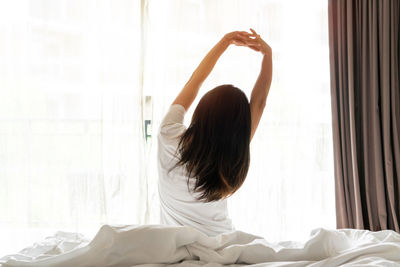 Rear view of woman with arms raised on bed at home