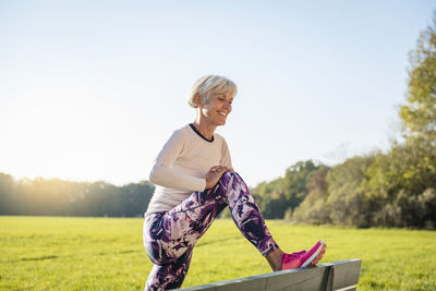 Smiling senior woman stretching on a bench in rural landscape