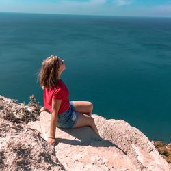 Side view of woman sitting on rock by sea during sunny day