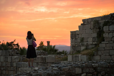 Rear view of woman standing on ruins in castle against orange sky