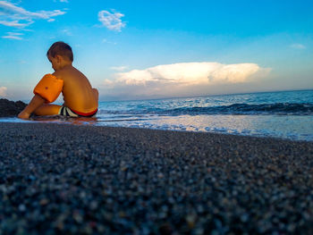 Surface level view of shirtless boy sitting at beach against blue sky