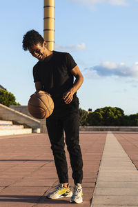 Smiling young man playing with basketball while standing on footpath during sunset