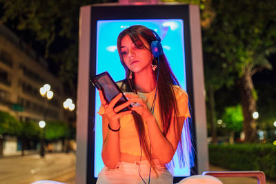 Interested female with beverage chatting on cellphone while listening to music from headset in night city
