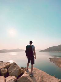 Rear view of man standing on rock looking at shore against sky