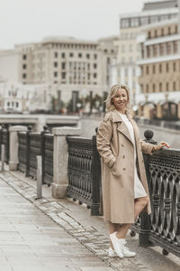 A middle-aged blonde woman with curly hair stands on the river embankment in the city.