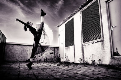 A ballerina in a black skirt does vertical splits on the roof of a building against the sky