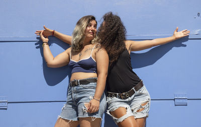 Two happy young women with arms raised against blue wall
