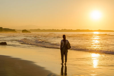Rear view of woman walking on shore at beach against sky during sunset